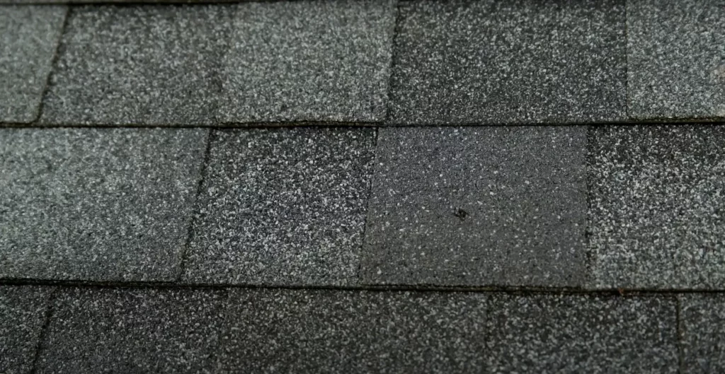 Close-up of dark gray asphalt roofing shingles with a textured, granular surface, showcasing the pattern and structure of traditional roofing materials.