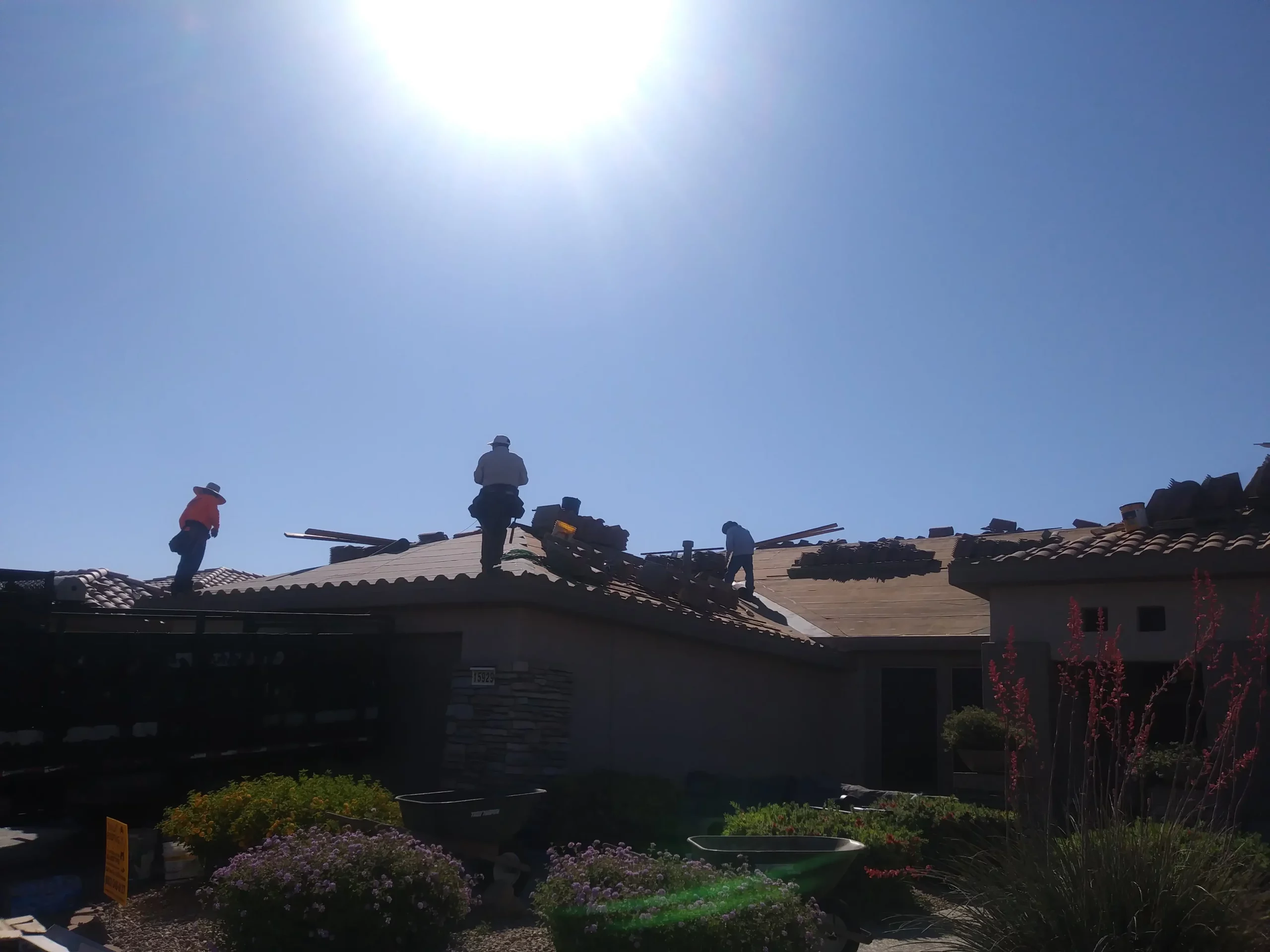 Roofing crew installing tiles on a sunlit Arizona home, silhouetted against a bright blue sky, working carefully along the roofline with tools and safety harnesses in place.