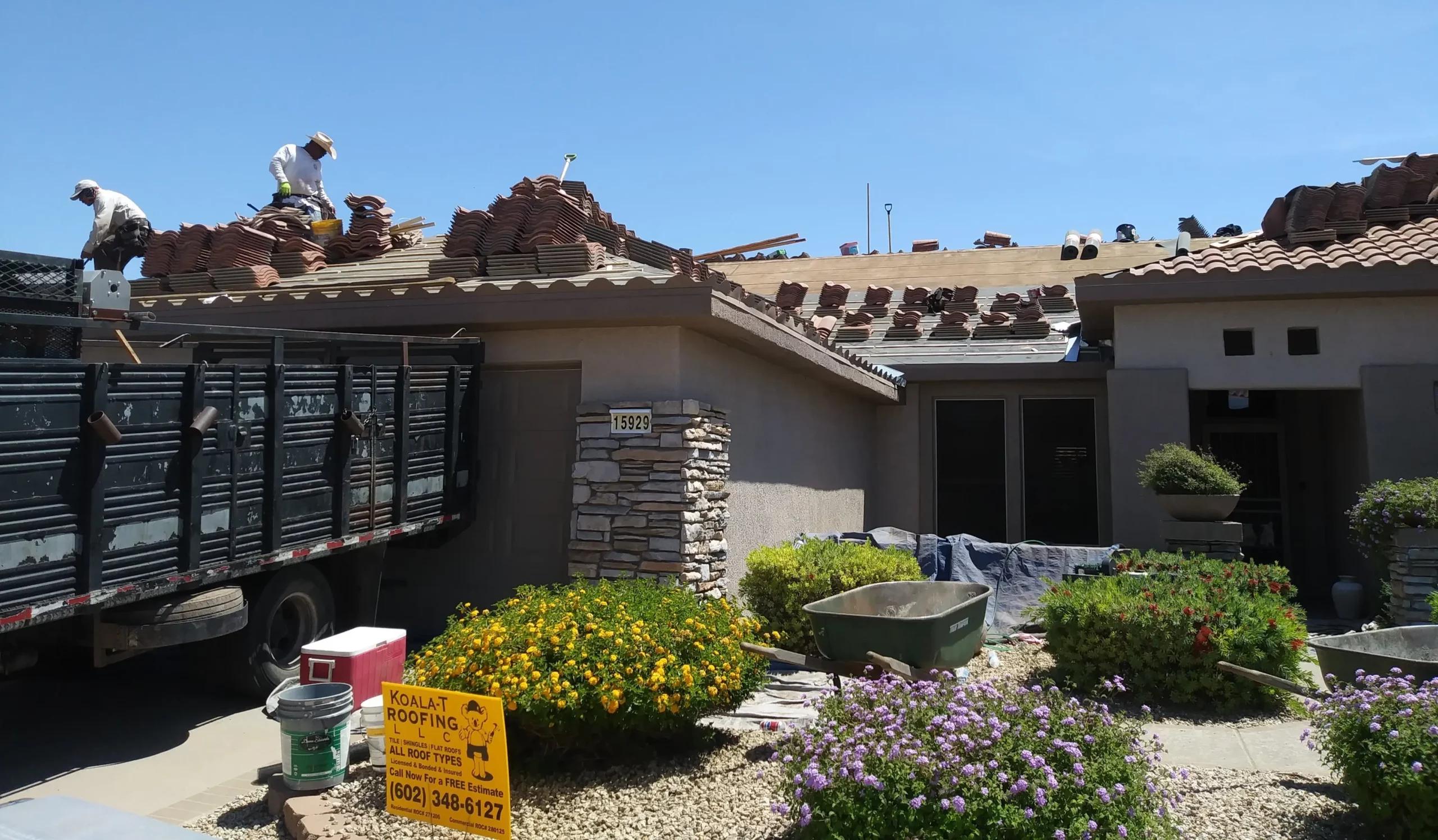 Two roofers working on a residential roof replacement project, surrounded by stacks of terracotta tiles and construction equipment in the front yard.