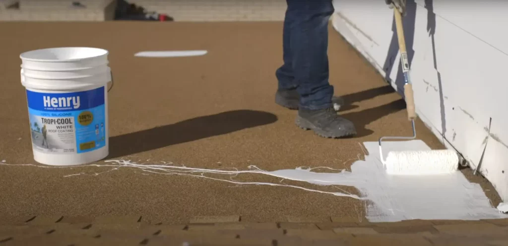 Worker applying white silicone roof coating using a paint roller to seal and protect a flat roof surface.