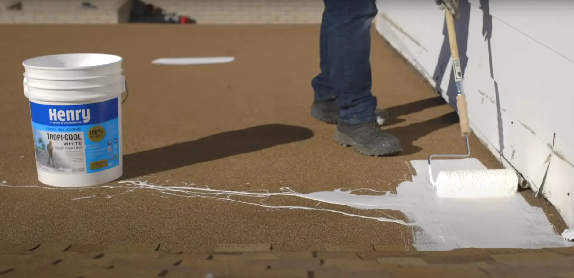 Worker applying white silicone roof coating using a paint roller to seal and protect a flat roof surface.