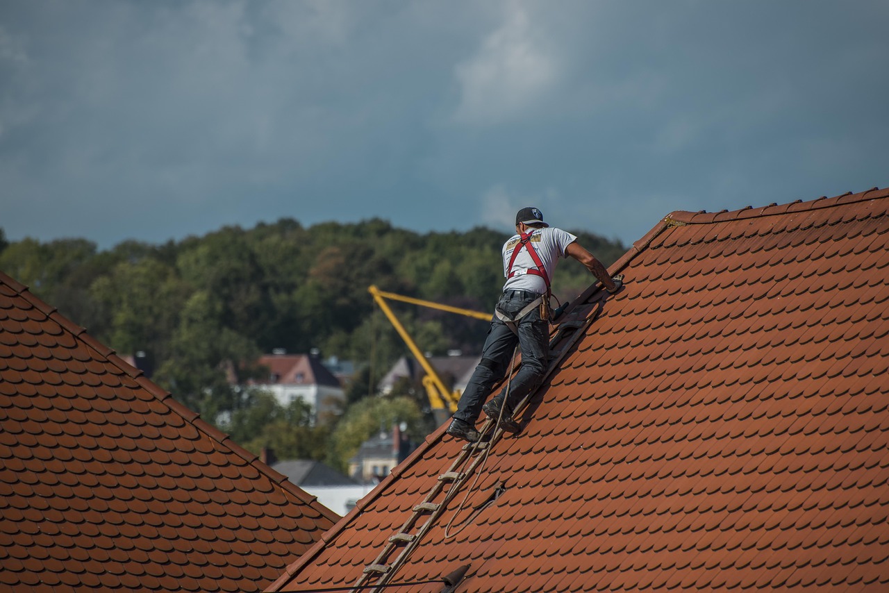 A roofer wearing a safety harness and climbing a ladder while inspecting a steep, red-tiled roof with a background of trees and distant houses.