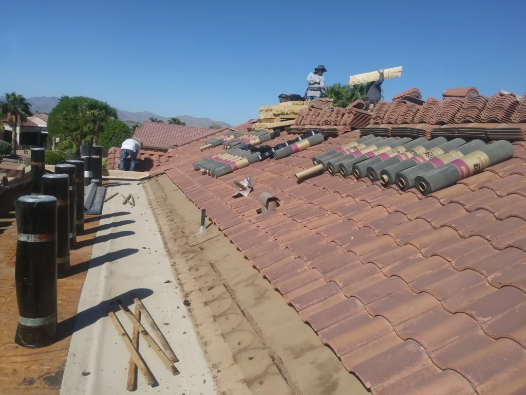 Roofers installing clay tiles on a residential rooftop under clear blue skies, with stacked materials and tools laid out for the ongoing roofing project.