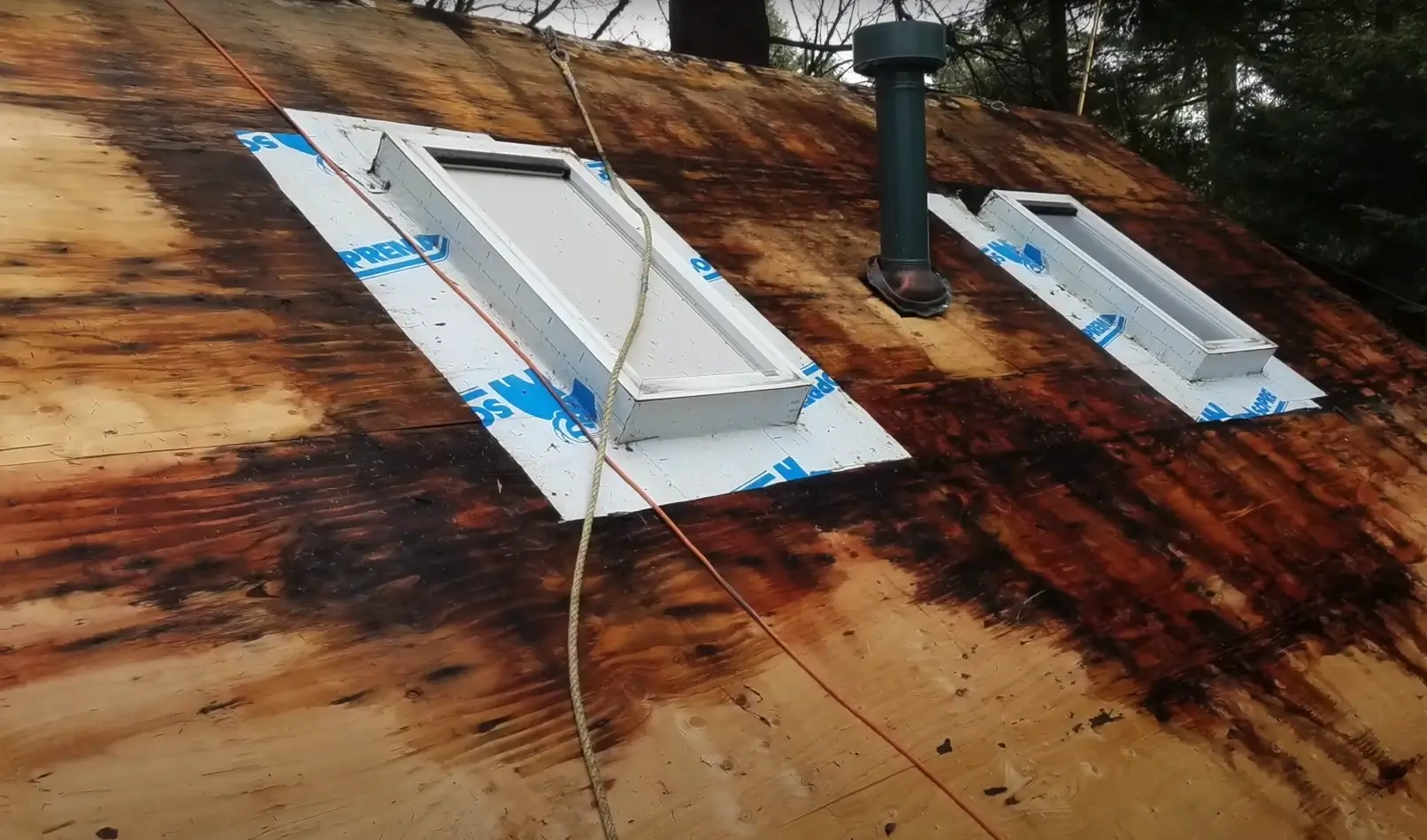 Partially exposed plywood roof with water stains and two freshly installed skylights sealed with waterproof flashing tape, indicating ongoing roof repairs in wet conditions.