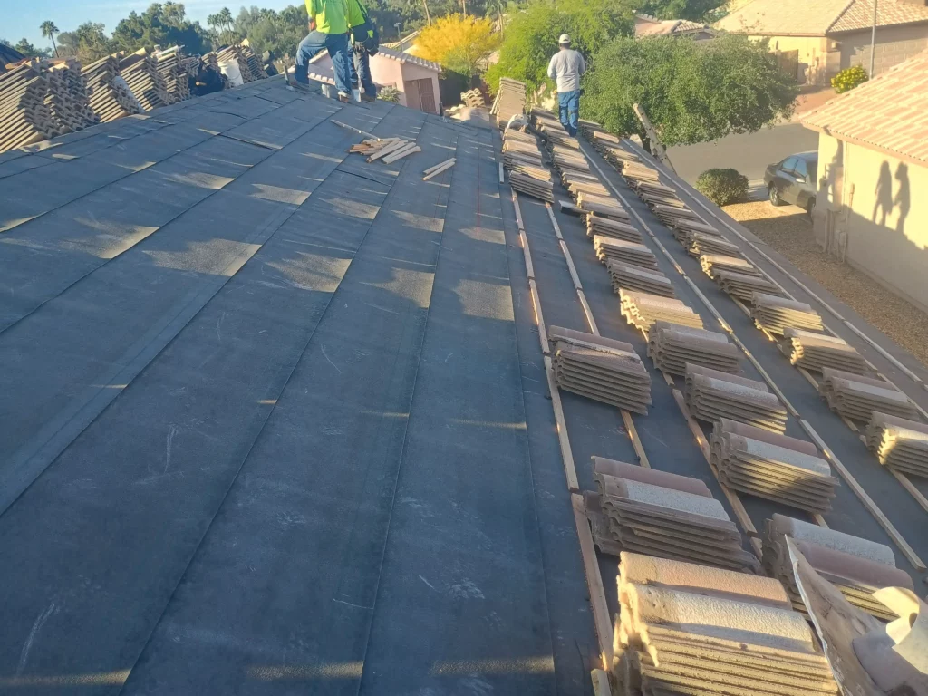 Stacks of clay roof tiles lined up on a home’s rooftop with underlayment installed and workers preparing for tile placement on a sunny residential street.