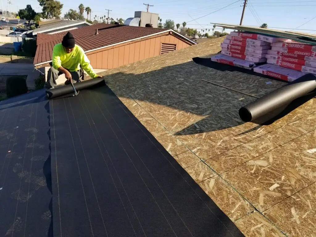 A roofing contractor in a neon yellow shirt unrolls black underlayment over a wooden roof deck, preparing for asphalt shingle installation on a sunny day in Arizona.