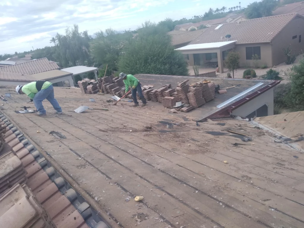 Two roofing contractors removing debris and inspecting an old, worn-out roof deck covered in remnants and damaged materials in Arizona.