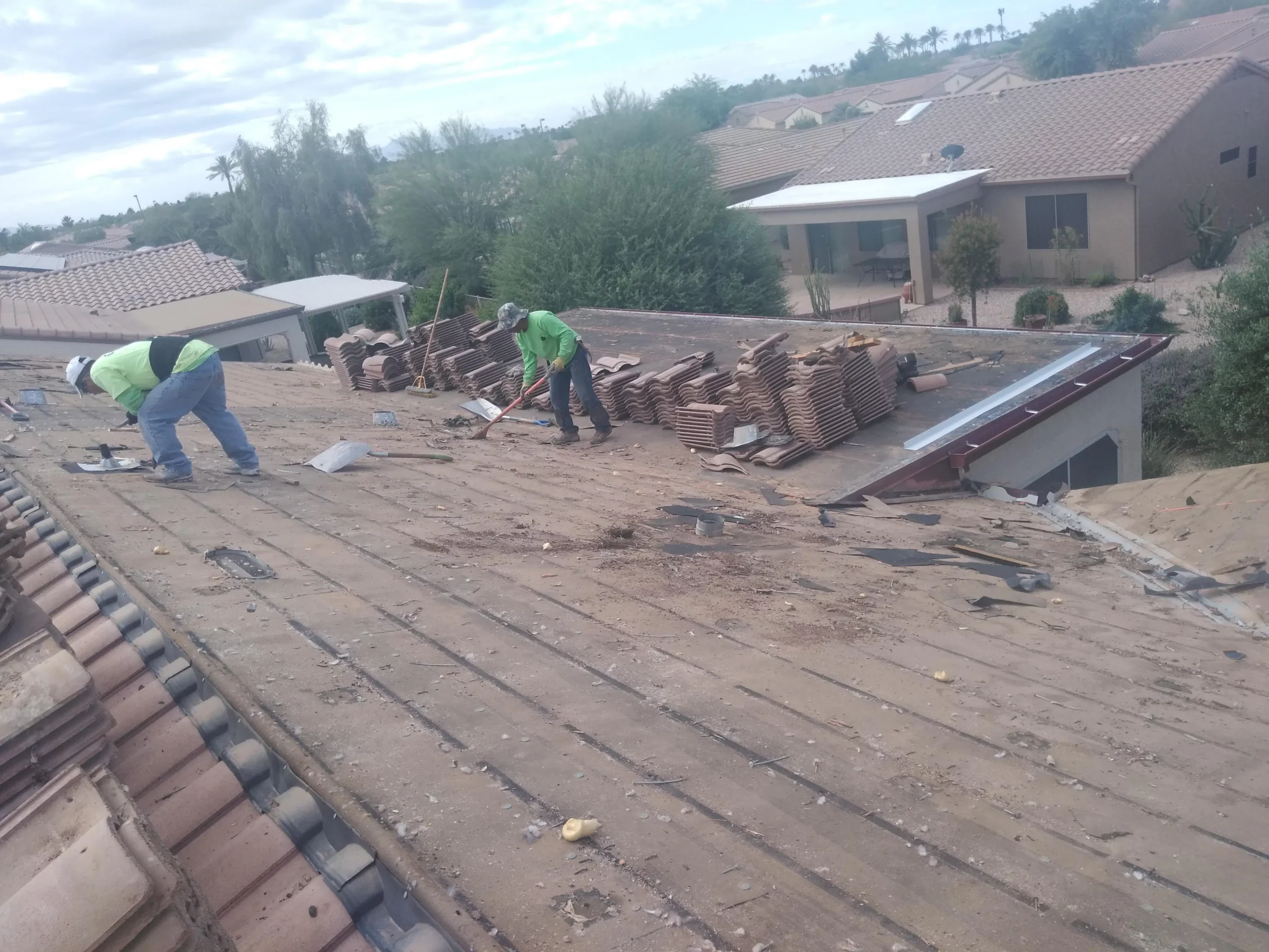 Two roofing contractors removing debris and inspecting an old, worn-out roof deck covered in remnants and damaged materials in Arizona.