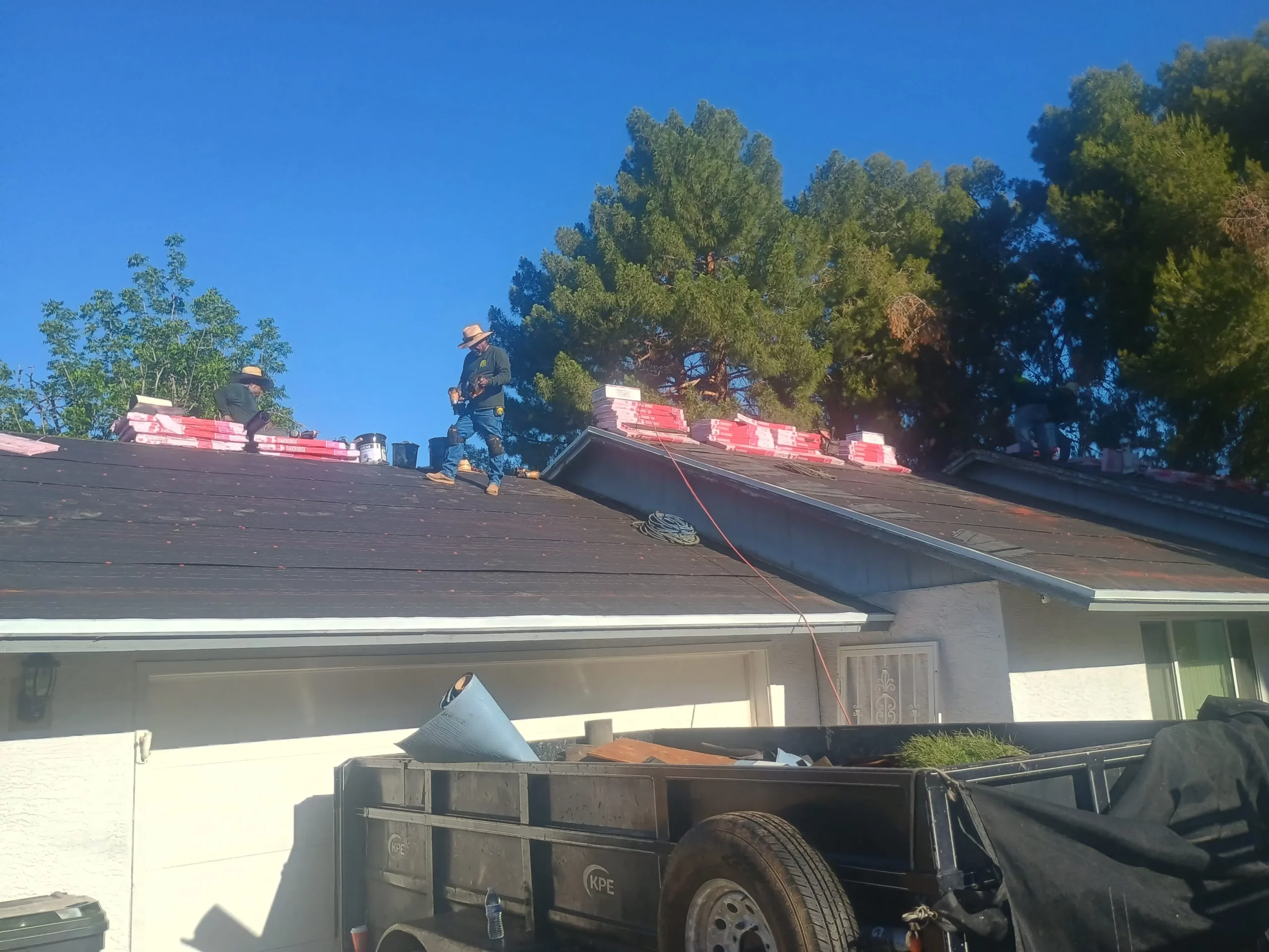 Arizona roofing crew standing on a residential roof deck covered in underlayment, surrounded by roofing material bundles and tools, with a debris trailer below.