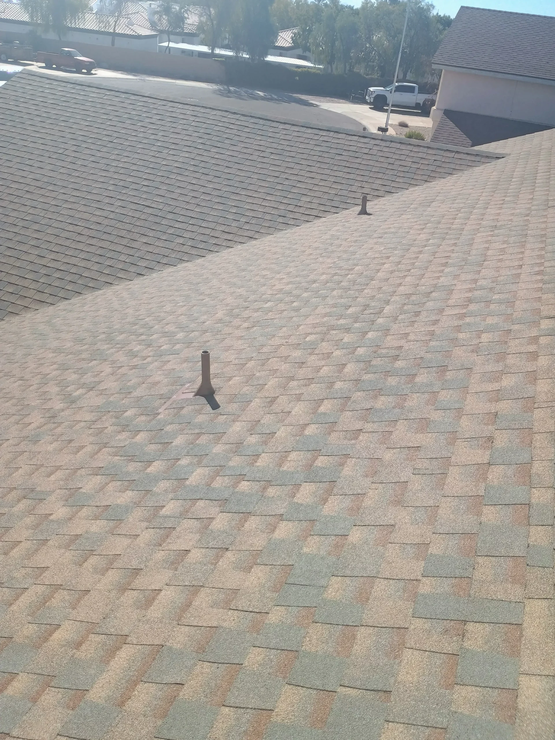 Close-up view of a clean, newly installed asphalt shingle roof with visible vent pipes under a clear blue sky in a residential neighborhood in Arizona.